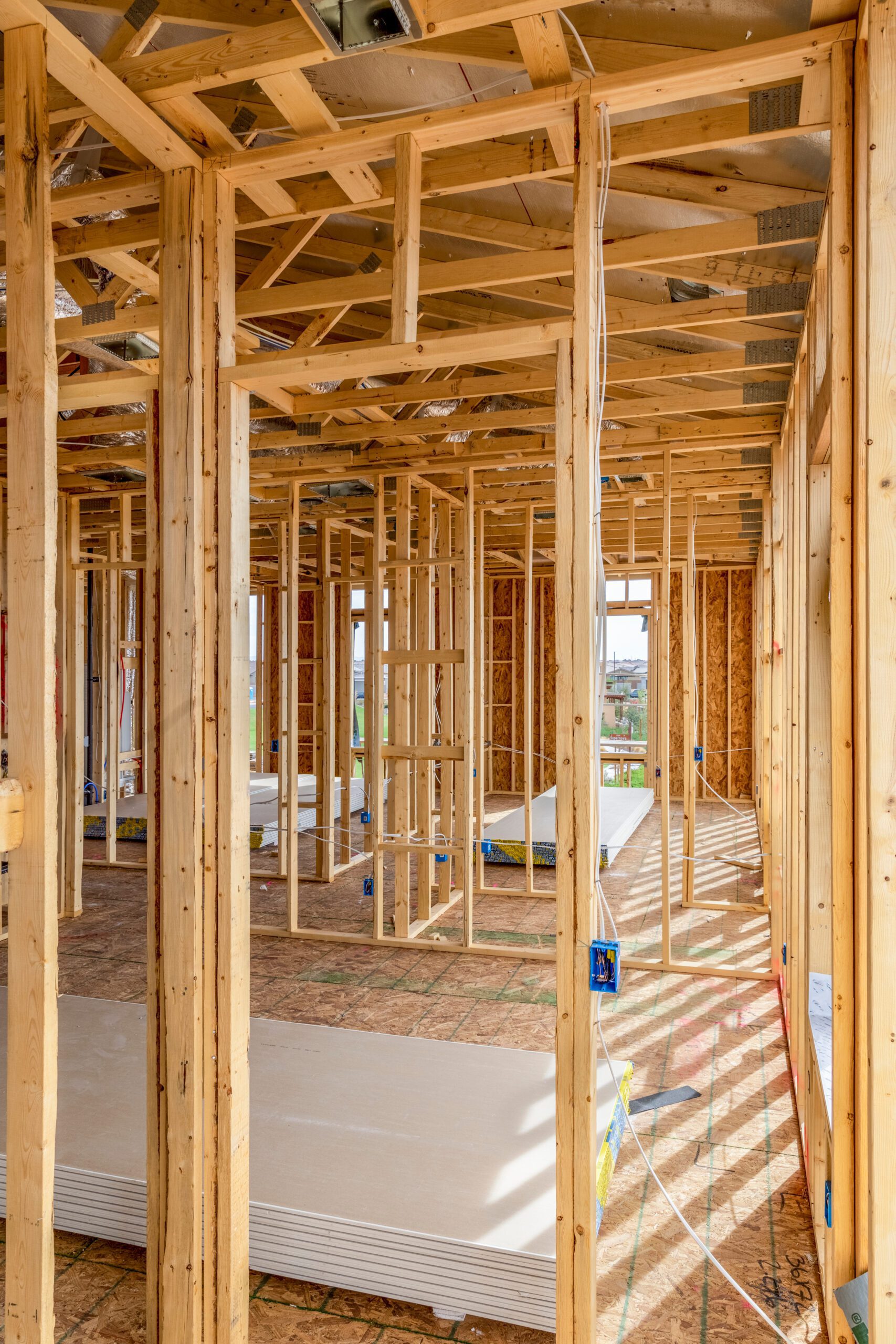 A partially constructed building shows wooden frames, drywall sheets on the floor, and electrical wiring running through the studs. Sunlight filters through open spaces, illuminating the construction site.